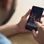 Over shoulder close up view of male analyst broker's hands holding touch screen device smartphone using tech ecommerce application of finance stock markets with graphs and numbers.