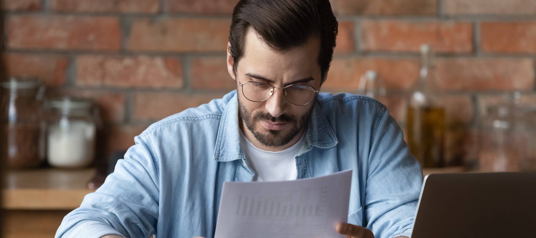 Serious distance employee analyzing statistics, data in paper financial reports at workplace with laptop.