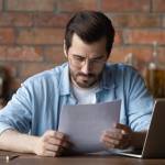Serious distance employee analyzing statistics, data in paper financial reports at workplace with laptop.