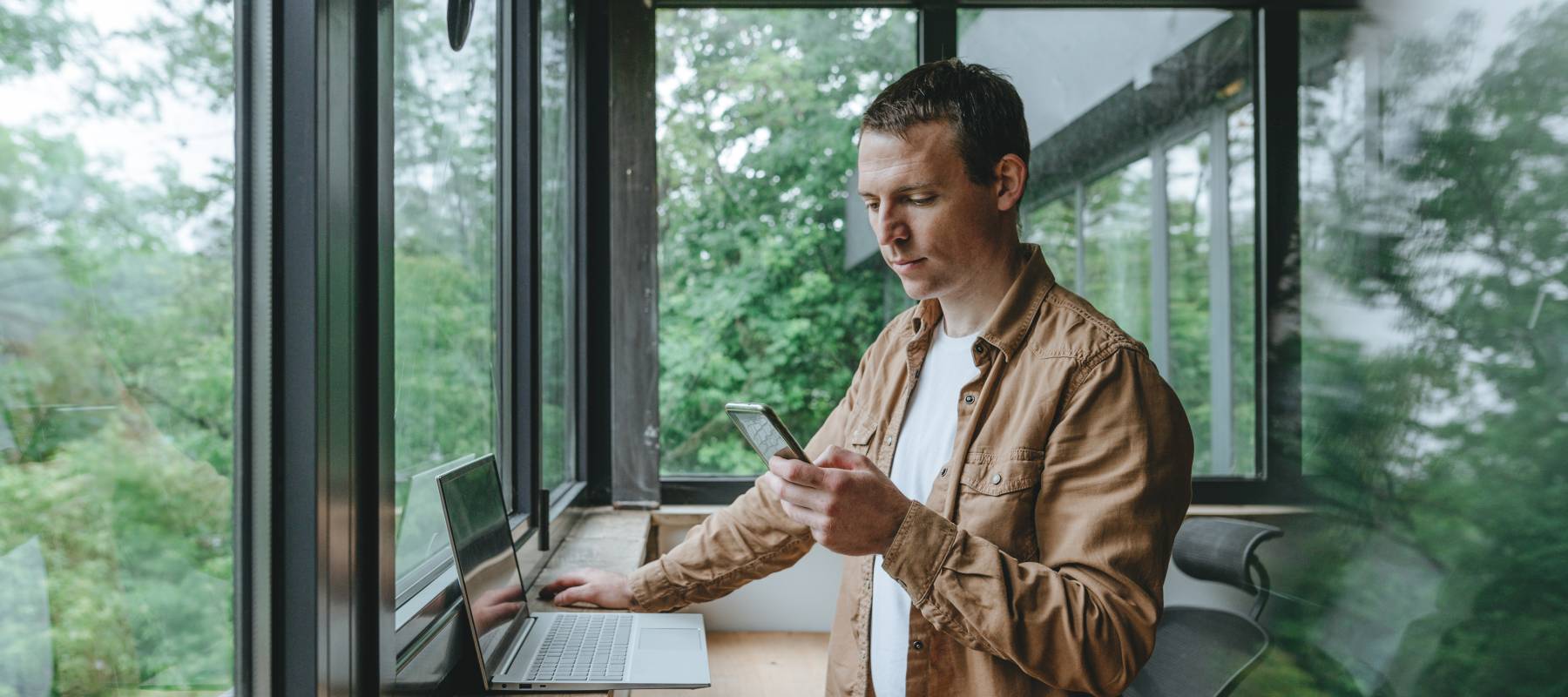 Young businessman in casual clothes standing in office near window looking at smartphone. Green modern workplace.