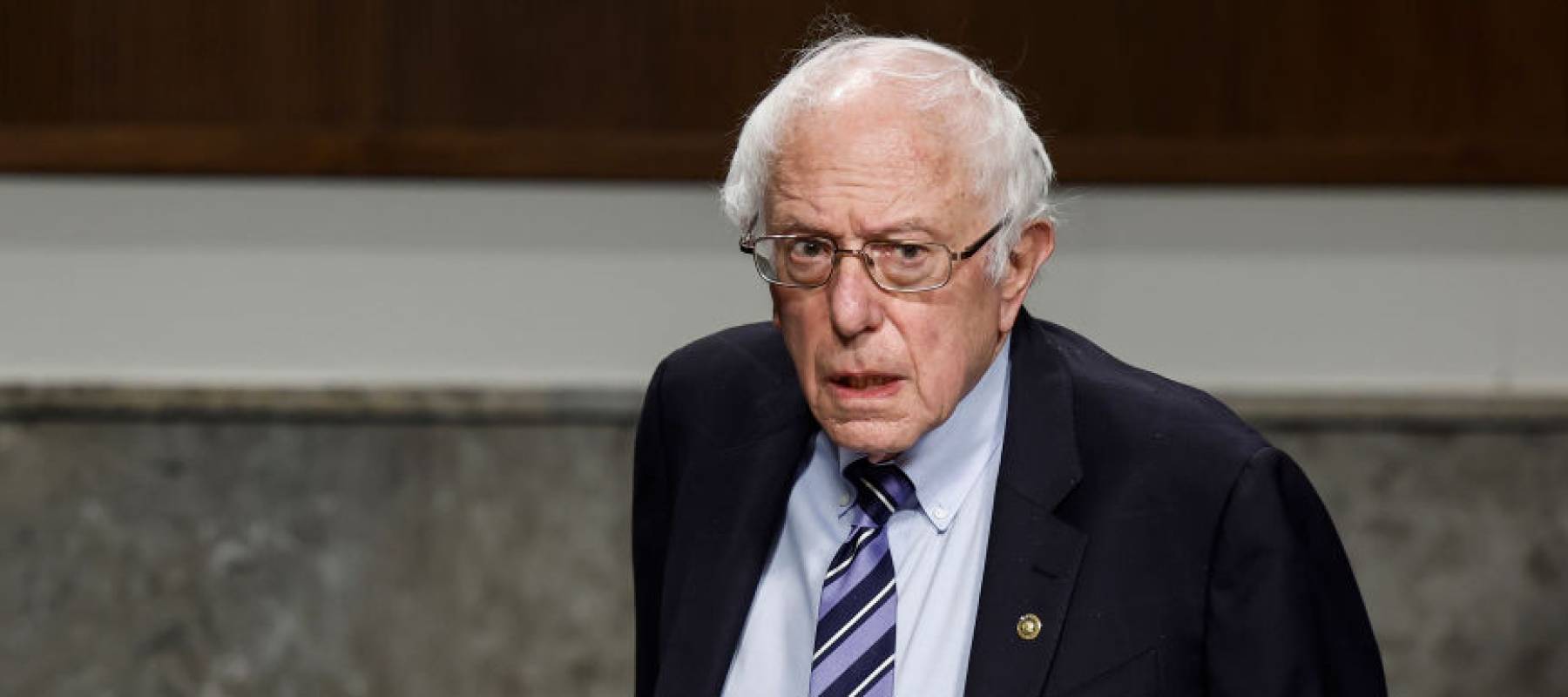 Bernie Sanders standing in Congress, looking serious and holding a binder.