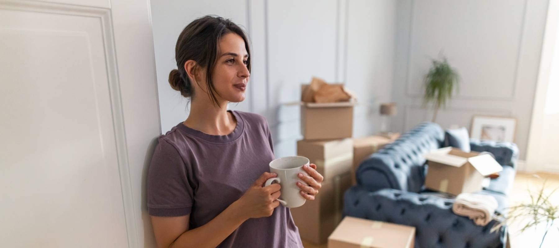 Young woman stands in doorway of apartment holding cup of coffee, boxes seen behind her.