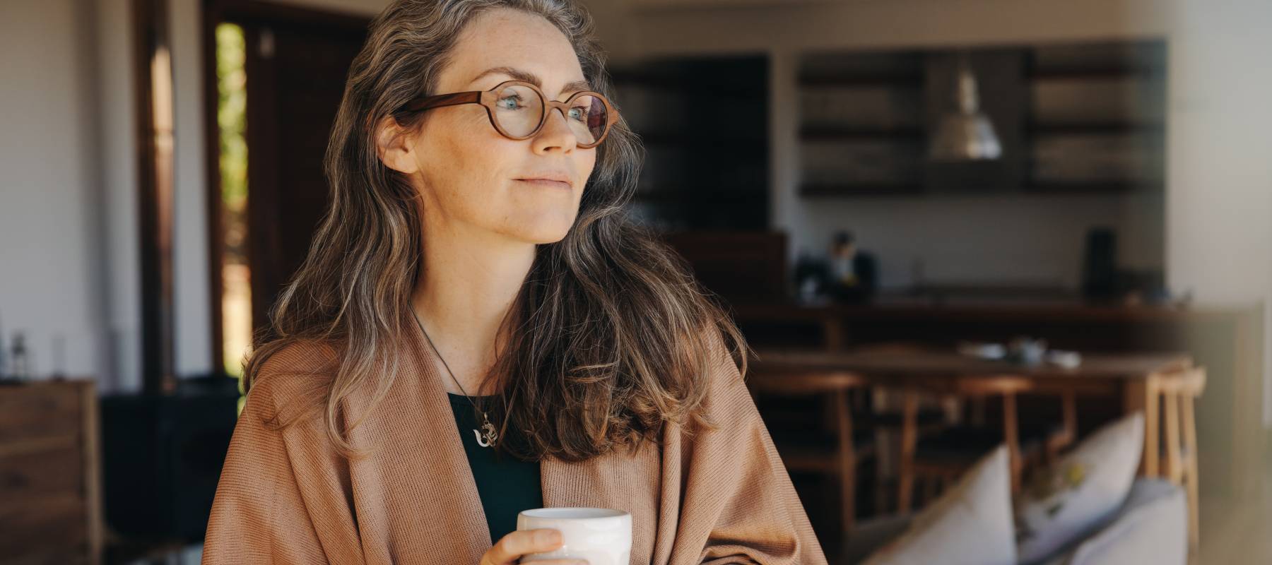 Senior woman looking away thoughtfully while standing with a cup of tea in her hands.