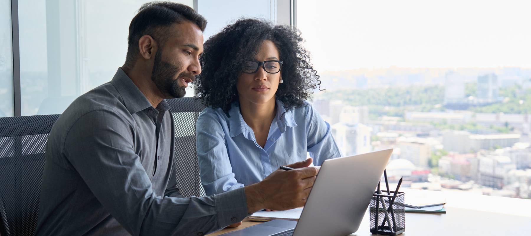 Indian male ceo executive manager mentor giving consultation on financial operations to female African American colleague intern using laptop sitting in modern office near panoramic window.