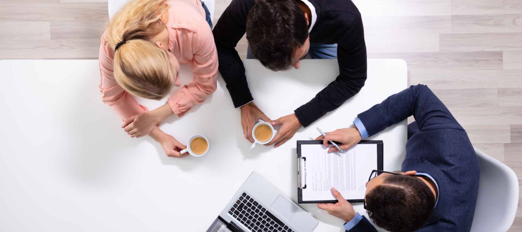 Happy Young Couple Consulting Male Friendly Financial Advisor With Coffee Cups At Office Desk