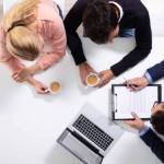 Happy Young Couple Consulting Male Friendly Financial Advisor With Coffee Cups At Office Desk