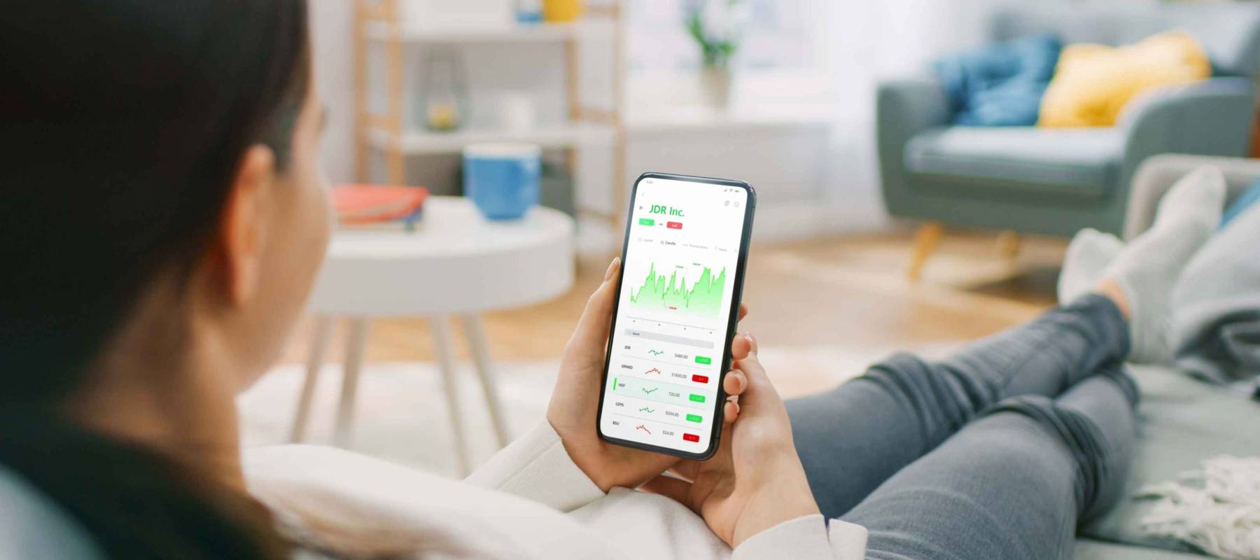Young Woman at Home Sitting on a Couch and Using Smartphone with Stock Market App to Check Share Prices. Graph is Going Up Making a Great Profit. In the Background Cozy Homely Atmosphere.