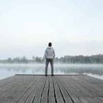 Young man standing alone on wooden footbridge and staring at lake.