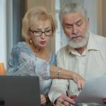 Senior aged couple checking accountancy and bills. Older retired couple holding computer and financial paper documents, checking domestic bills, discussing expenses together at home.