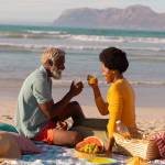 African american couple toasting juice while sitting with food on blanket against sea at sunset. retirement