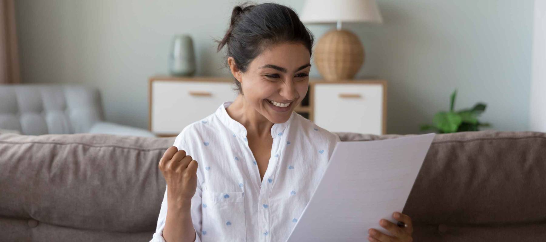 Excited cheerful young Indian woman getting good news letter, reading paper document at home, making winner yes hand, feeling happy, smiling, laughing, celebrating, success, achieve