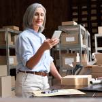 Female mature senior small business owner using mobile app checking parcel box.