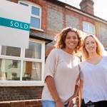 Portrait Of Two Women Standing Outside New Home With Sold Sign