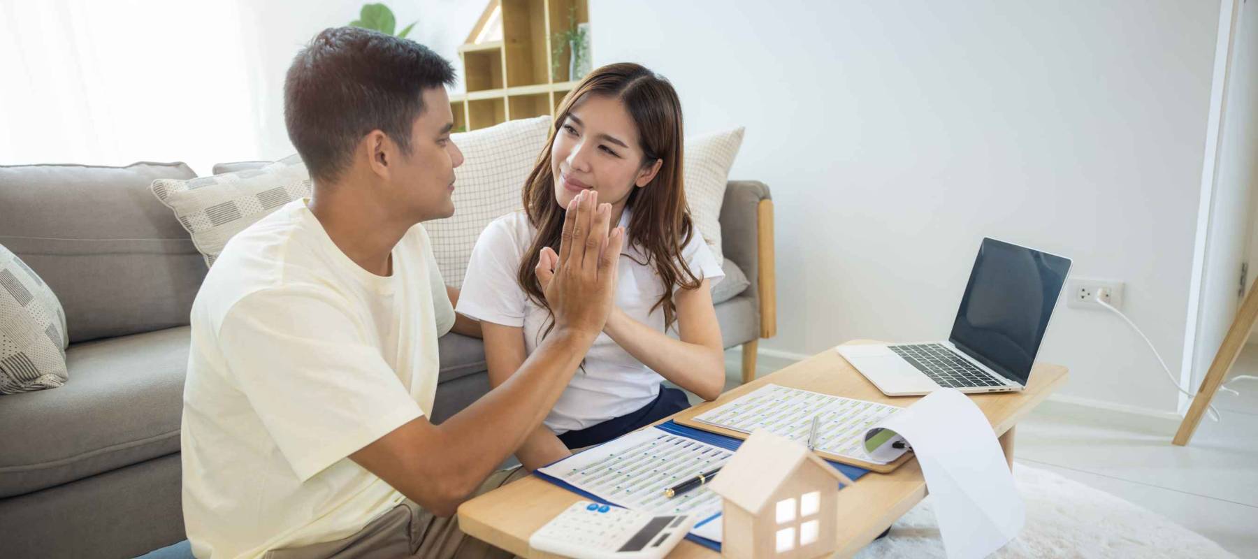 Asian couple in home high-fiving by coffee table with calculators, paperwork and laptop