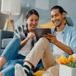 Happy asian  couple man and woman looking at tablet together seated on floor in front of couch