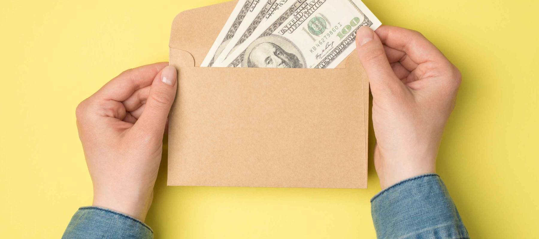 First person top view photo of female hands holding open craft paper envelope with hundred dollars banknotes on isolated yellow background with copyspace