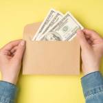 First person top view photo of female hands holding open craft paper envelope with hundred dollars banknotes on isolated yellow background with copyspace