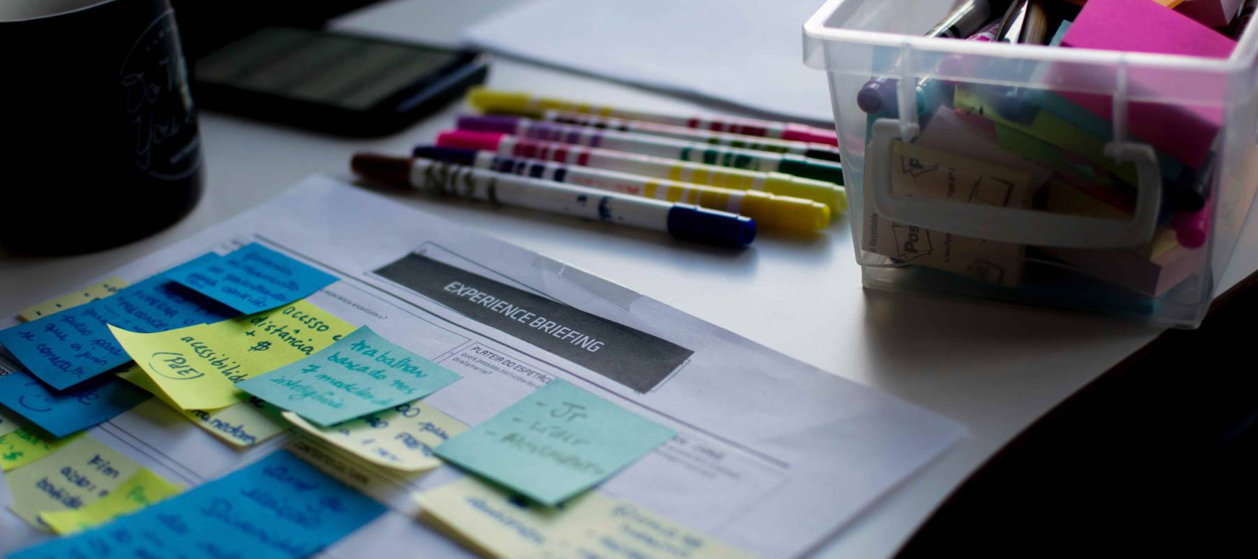 sticky notes on paper beside coffee mug, markers of various colors and a bin of more markers and sticky notes