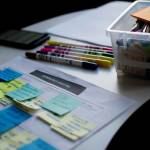 sticky notes on paper beside coffee mug, markers of various colors and a bin of more markers and sticky notes