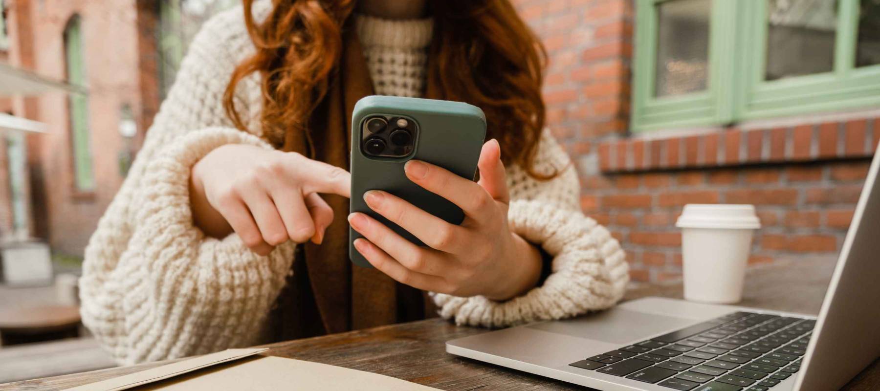 Close up of redhead girl using smartphone and working on laptop while sitting in cafe outdoors