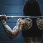 rear picture of tattoed woman's back at gym holding barbell with weights standing ready to do a squat to get strong