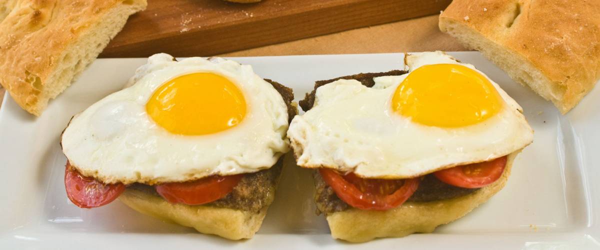 Scrapple and sunny side up eggs on a split hoagie roll with extra bread on a wooden cutting board in background