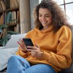 Happy millennial hispanic teen girl checking social media holding smartphone at home.