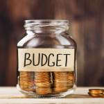 Glass jar with money and a sticker with the words BUDGET, on a wooden table.