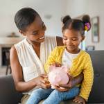 Smiling mature african american mother helping daughter sitting on lap putting money in piggy bank. Cute little black girl saving money by adding a coin in piggy bank with mother at home.