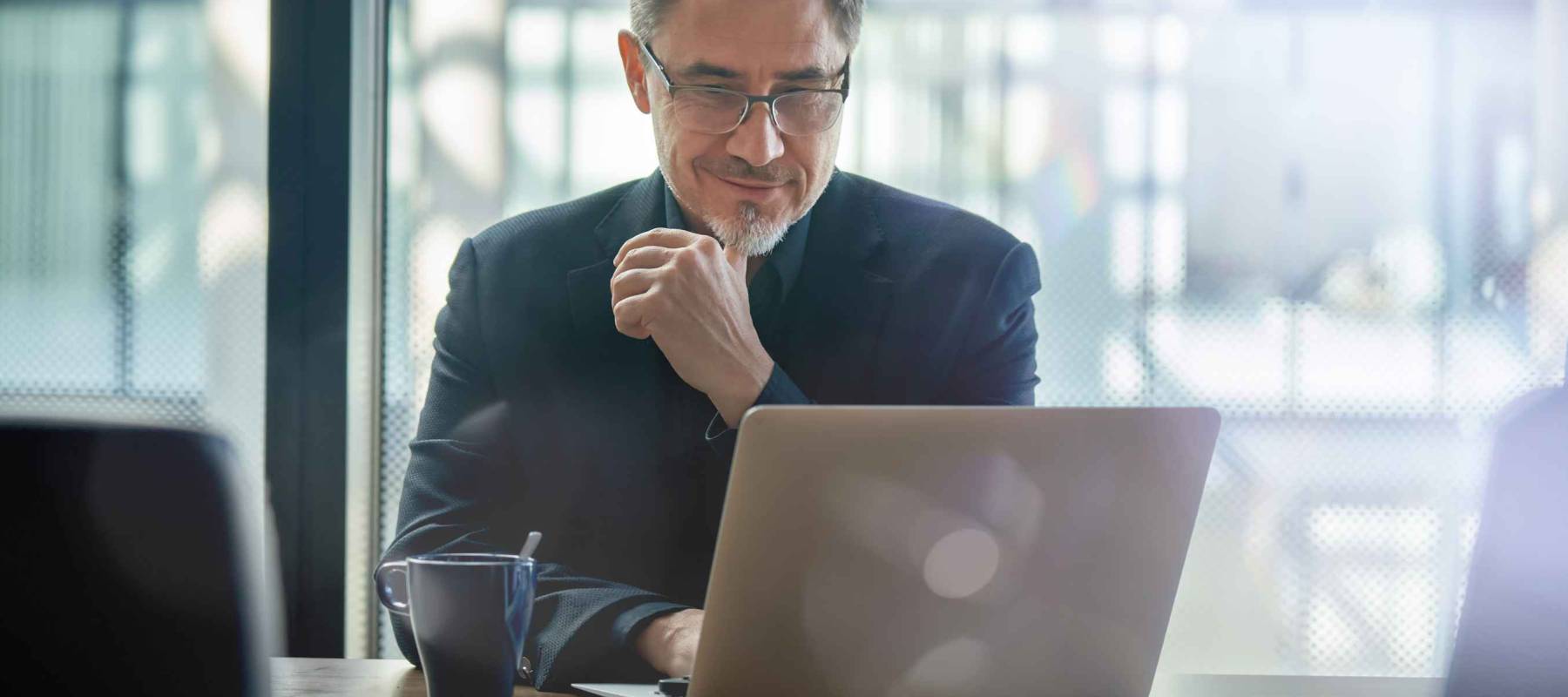 Business portrait - businessman sitting in in office working with laptop computer. Mature age, middle age, mid adult man in 50s with happy confident smile. Copy space.