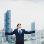 Happy Asian male engineer or businessman Wearing a yellow helmet is standing on the rooftop of building and a skyscraper over a cityscape