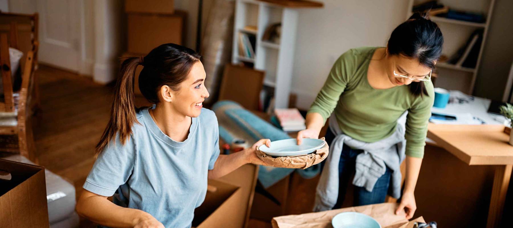 Happy woman and her Asian female friend wrapping fragile things into a paper while preparing to move out of the apartment.