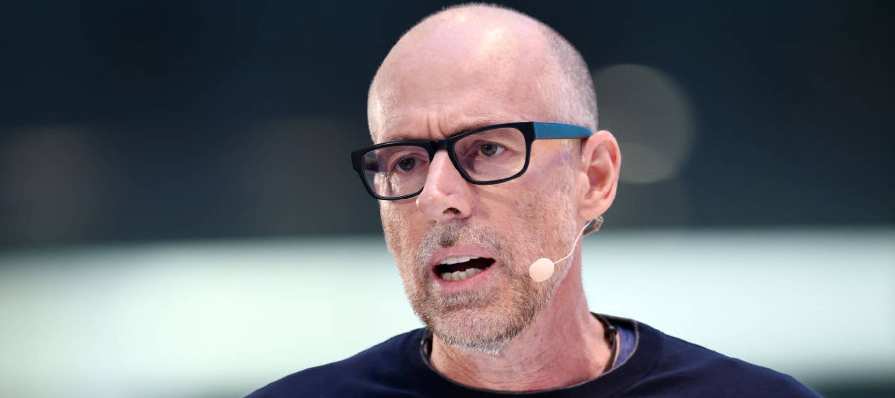 Professor Scott Galloway talking on stage wearing black glasses and a microphone.