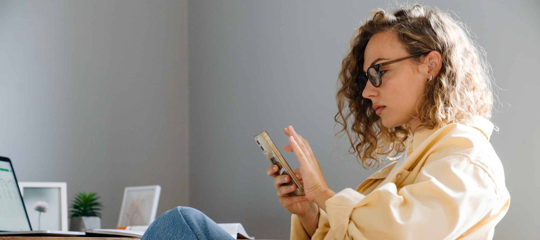 Young curly serious woman in glasses with phone sitting by table in cozy room at home