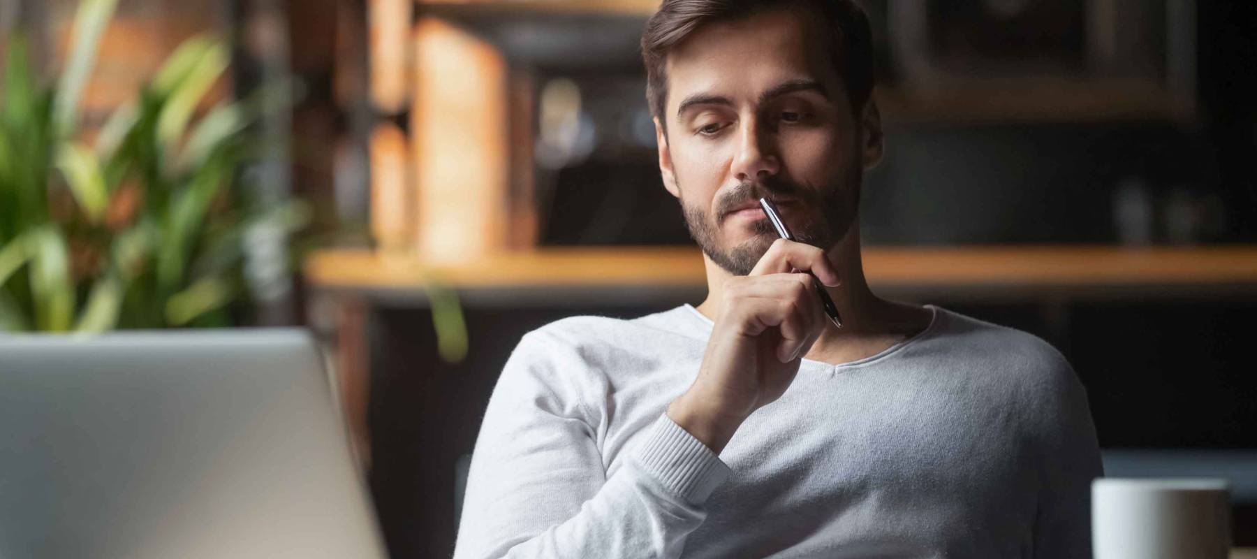 Pensive bearded man sitting at table drink coffee work at laptop thinking of problem solution, thoughtful male employee pondering considering idea looking at computer screen making decision