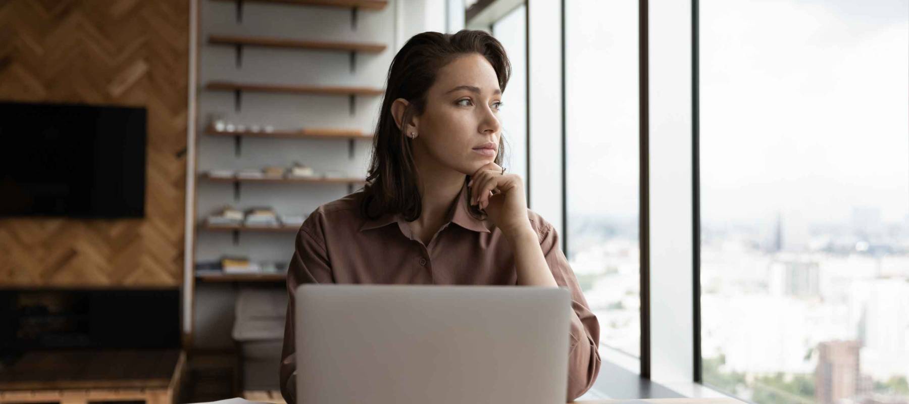 Thoughtful business owner woman looking out of window at city, sitting at workplace with laptop, thinking over solutions, challenges, company future vision, making decision, feeling uncertain