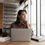 Thoughtful business owner woman looking out of window at city, sitting at workplace with laptop, thinking over solutions, challenges, company future vision, making decision, feeling uncertain