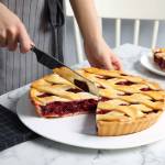 Woman cutting tasty cherry pie at white marble table, closeup