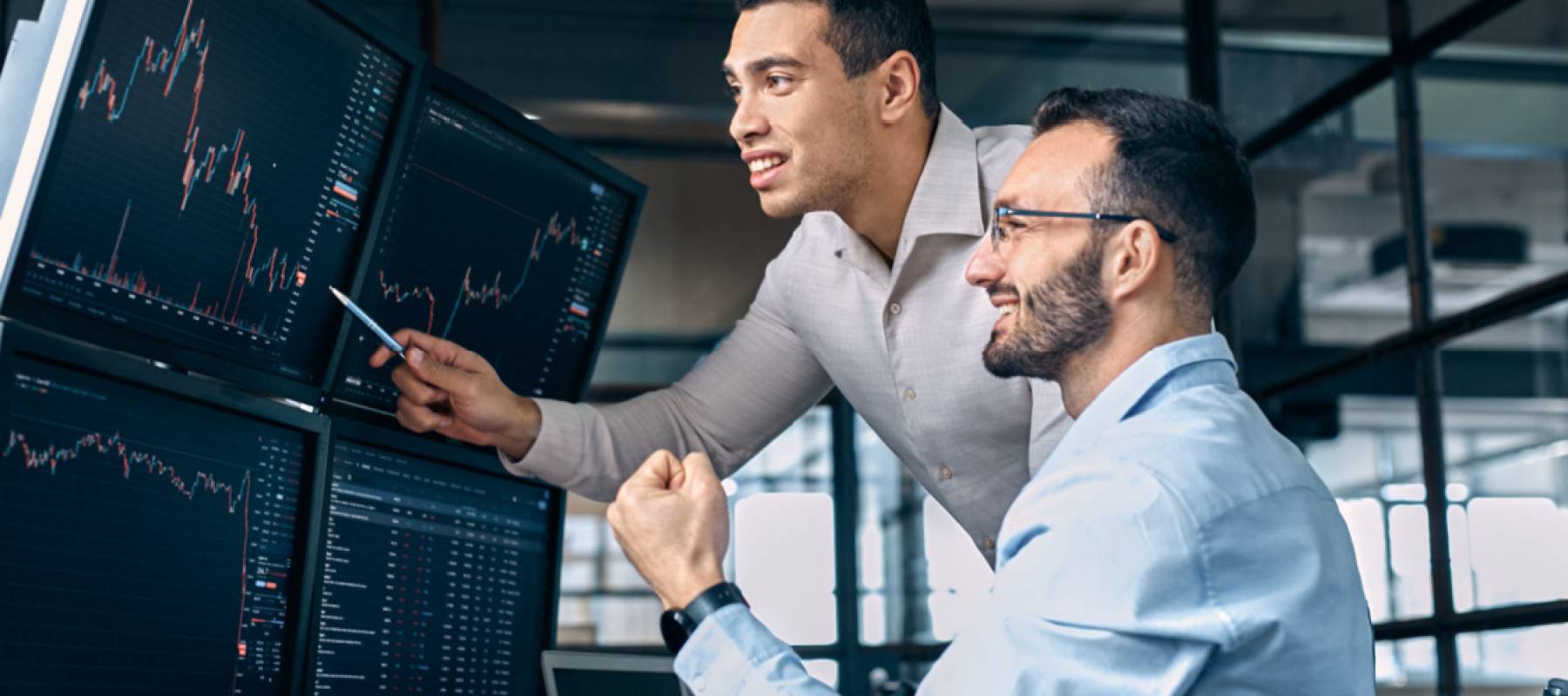 Two men traders sitting at desk at office together monitoring stocks data candle charts on screen analyzing price flow smiling cheerful having profit teamwork concept