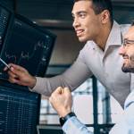 Two men traders sitting at desk at office together monitoring stocks data candle charts on screen analyzing price flow smiling cheerful having profit teamwork concept