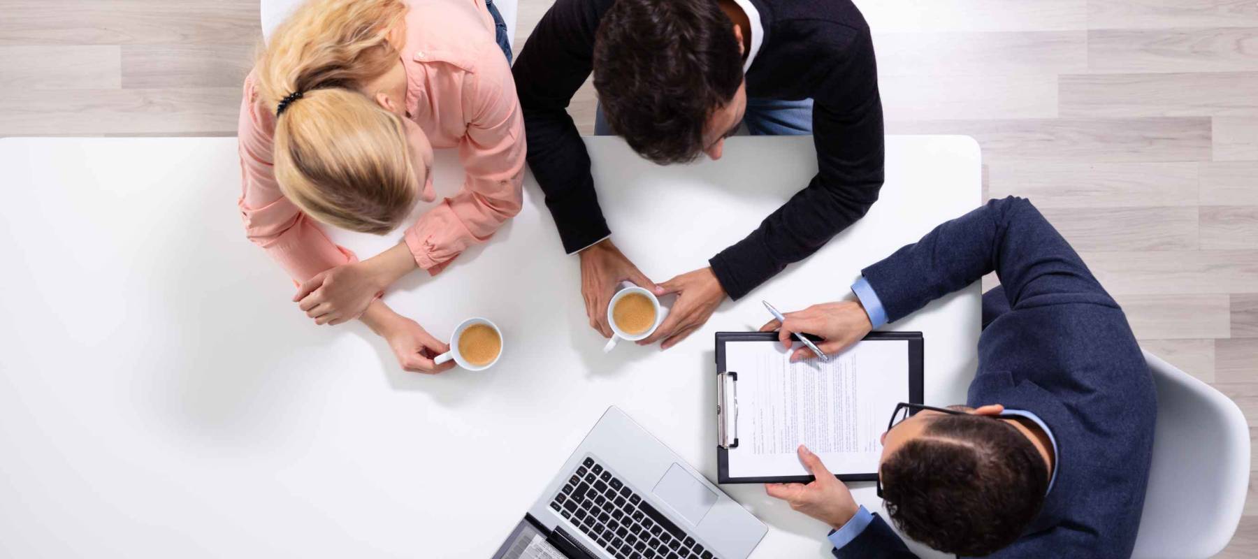 Happy Young Couple Consulting Male Friendly Financial Advisor With Coffee Cups At Office Desk
