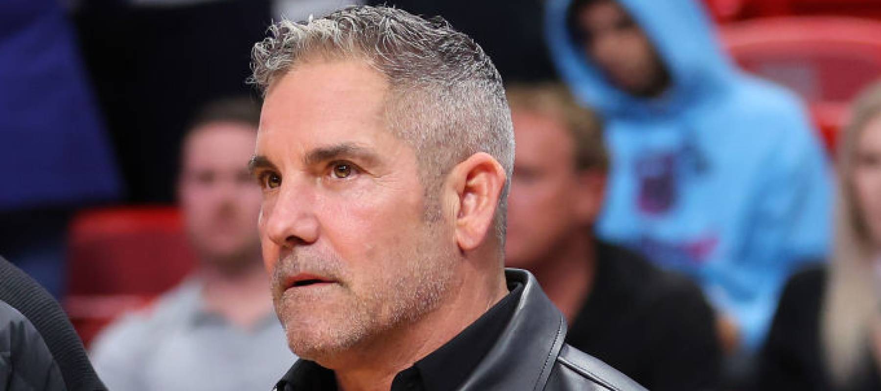 Grant Cardone sits in the front row at a basketball game, looking serious and leaning forward.