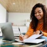 Smiling young black woman with open laptop writing notes happy with her dividends