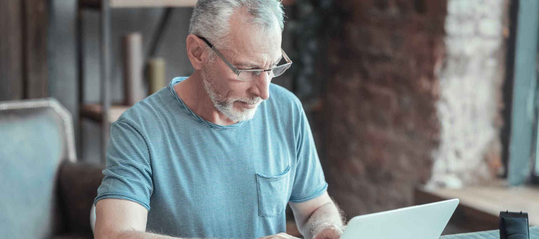 Full concentration. Serious bespectacled aged man sitting in the room by the table having important task and working with the laptop.