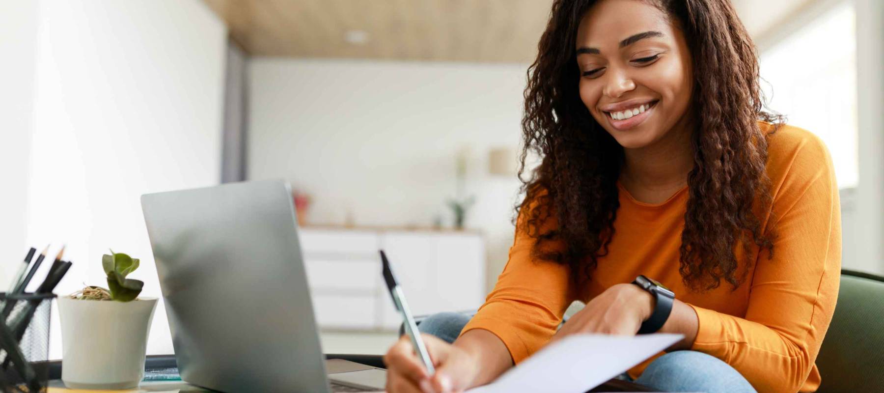 Business And Education Concept. Smiling young black woman sitting at desk working on laptop writing letter in paper notebook, free copy space. Happy millennial female studying using pc