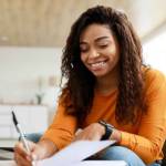 Business And Education Concept. Smiling young black woman sitting at desk working on laptop writing letter in paper notebook, free copy space. Happy millennial female studying using pc