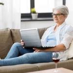 technology, old age and people concept - happy senior woman in glasses with laptop computer and glass of wine at home