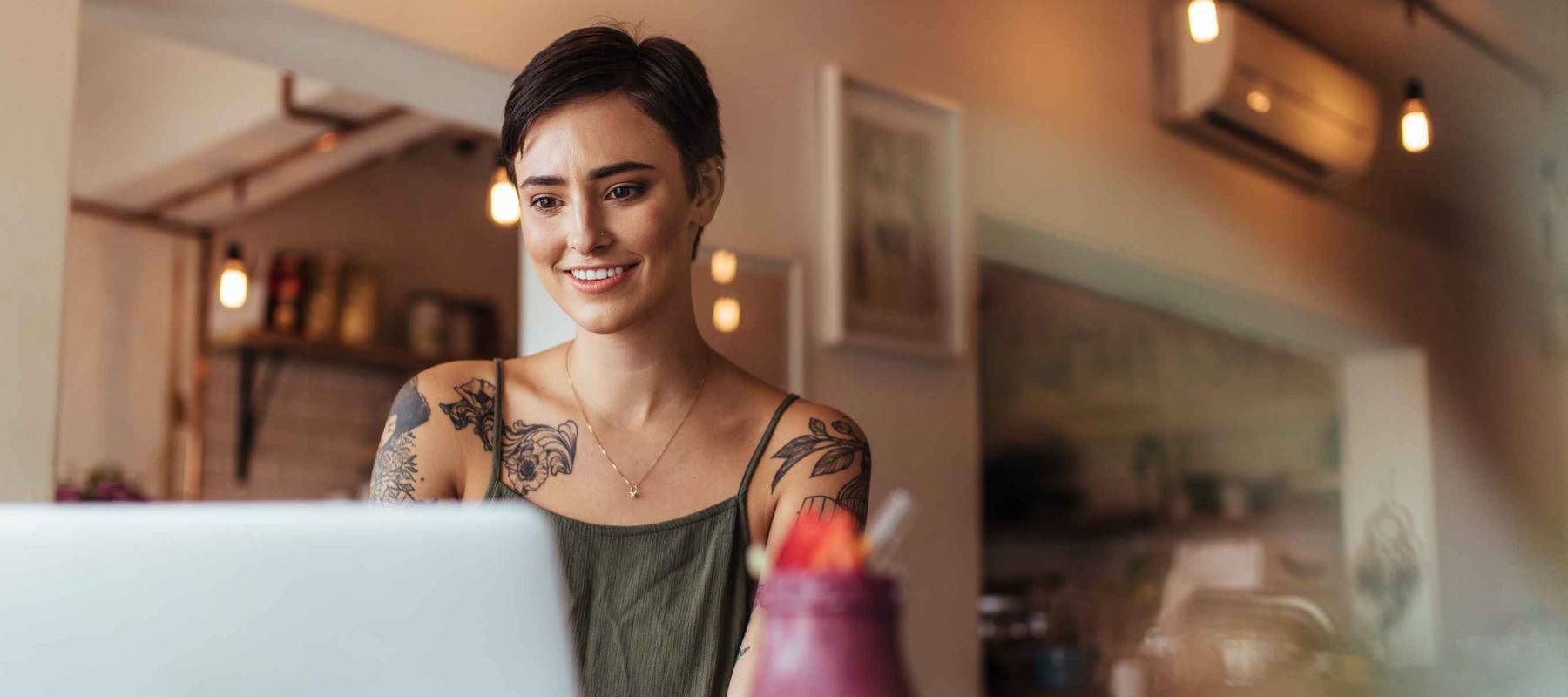 Woman entrepreneur working on laptop computer at home with a smoothie on the table. Woman with tattoos on her body working on laptop.