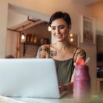 Woman entrepreneur working on laptop computer at home with a smoothie on the table. Woman with tattoos on her body working on laptop.