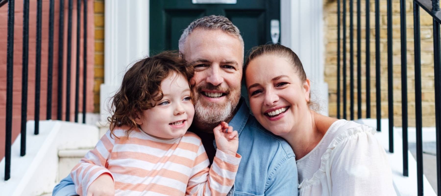 Happy family of three sit on front porch of house, smiling for camera.